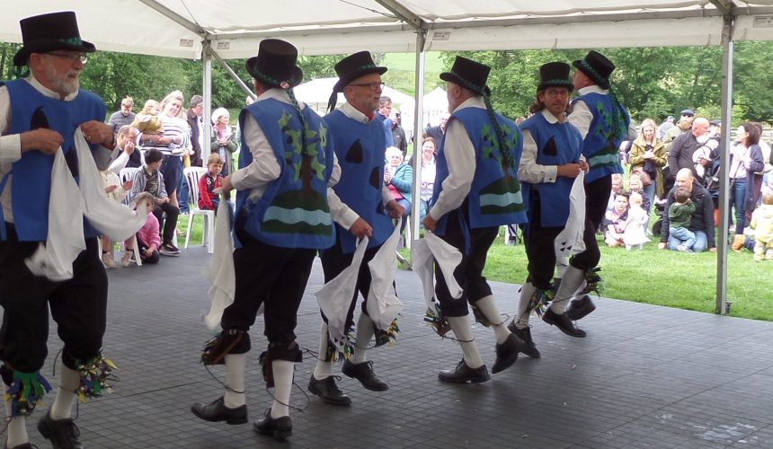 Morris dancers dance in a line. They wear blue waistcoats