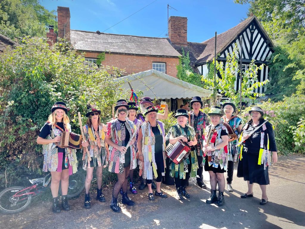 Morris dancers pose for a photograph. They wear multi coloured tatter jackets and black hats.