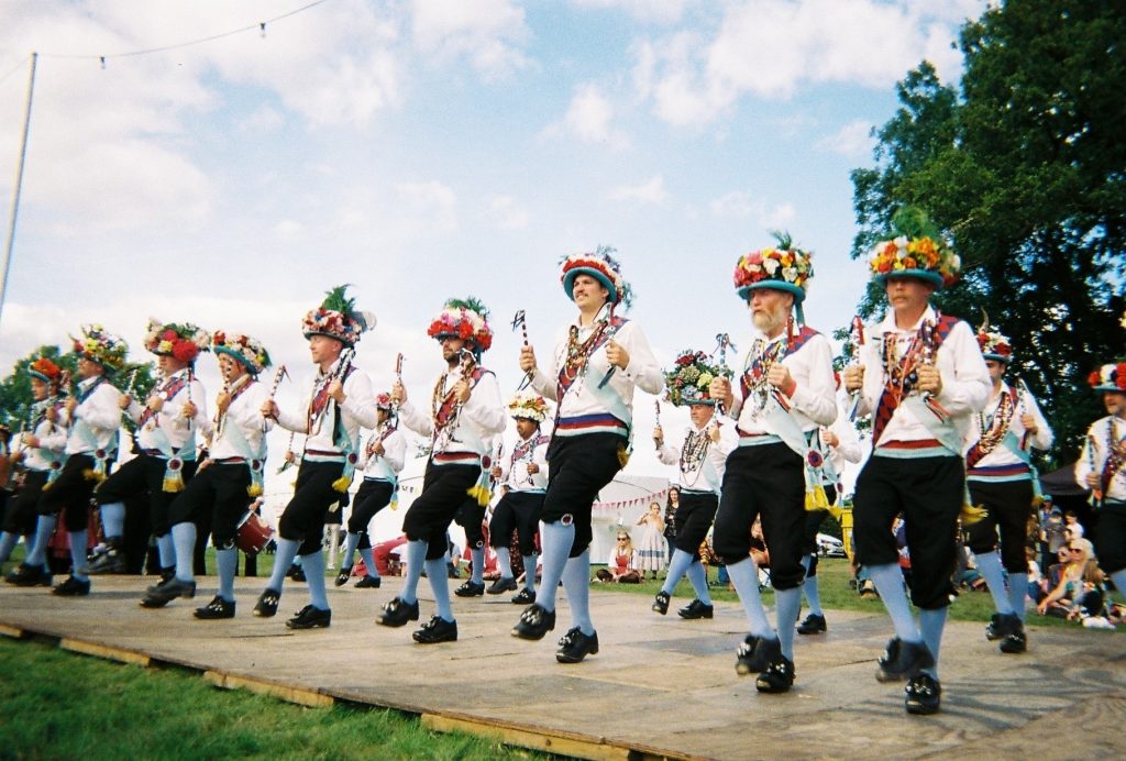 Lots of morris dancers are dancing in two lines. They wear clogs, sky blue socks, and sashes of blue and burgundy. They are wearing blue bowlers hats decorated with flowers and foliage.
