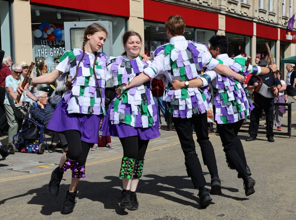 Four young morris dancers hold each other at the waist and dance in a line. They are wearing tatter jackets of purple, greens and white.