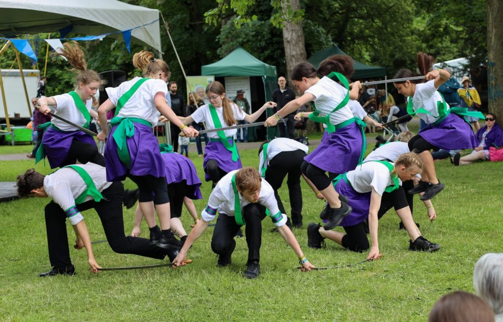 Young morris dancers are standing in a circle, holding longswords. Some are kneeling outwards with their swords on the ground, and some are jumping inwards over the swords. They are wearing purple, white and green.