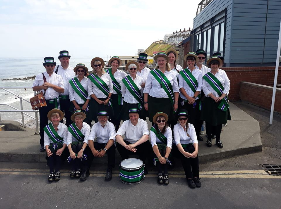 A group of morris dancers post for a photo. They are wearing white shirts and green and purple sashes, and straw boaters.