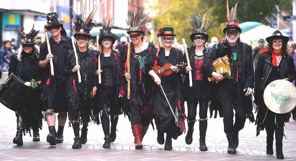 Nine morris dancers walk towards the camera in a line. They wear black clothing with flashes of red and green, and hats decorated with feathers. Some are holding sticks, and some are holding instruments.