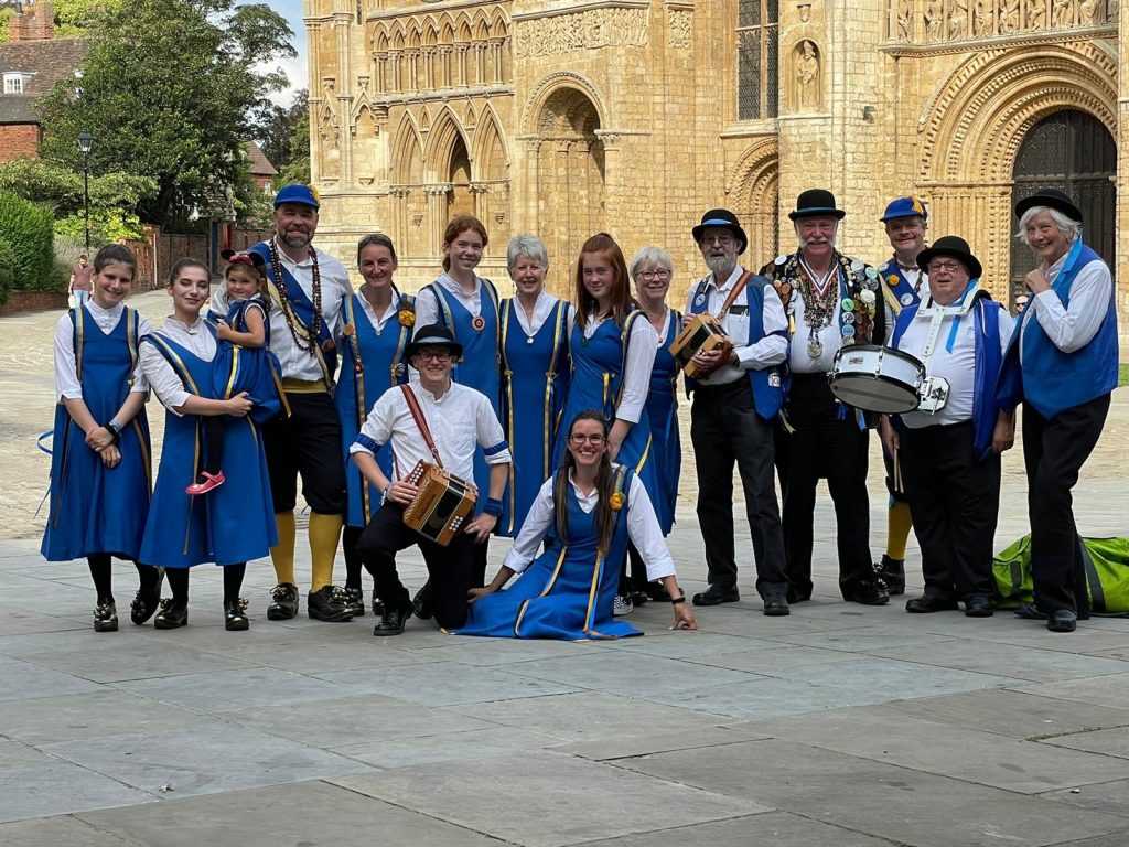 A group of morris dancers post for a photograph. The dancers wear blue dresses or black breeches, yellow socks, and blue sashes. The musicians wear blue waistcoats and carry instruments.