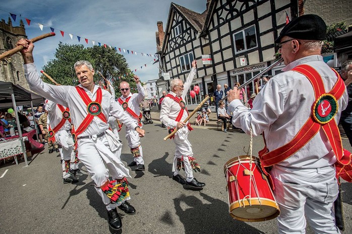 Dancers in white shirts and red baldricks raise their arms in the air as a musician plays the pipe and tabor