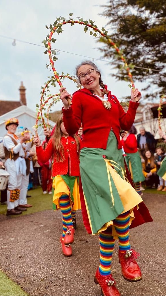 A Chelmsford Morris Dancer holds a garland above her head and smiles at the camera.